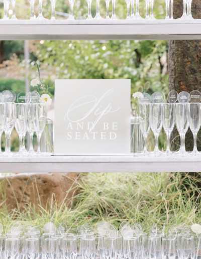 Shelves filled with arranged champagne flutes, with a sign reading "Sip and Be Seated," surrounded by flowers and greenery.